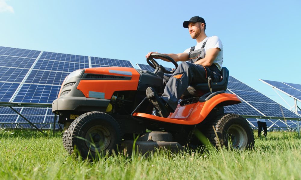 A man working at solar power station. A worker on a garden tractor mows grass on a solar panel farm.